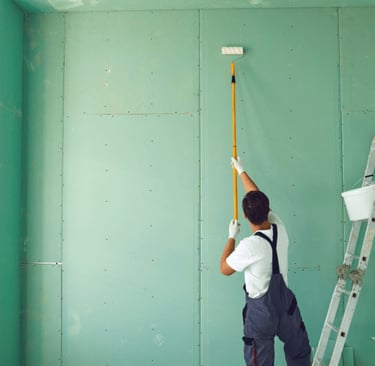 Professional painter in overalls using a long roller to prime green drywall panels during interior renovation.