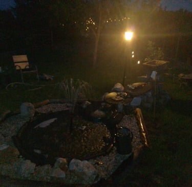 A dark nighttime scene showing a garden fountain illuminated by a warm lamp beside it,