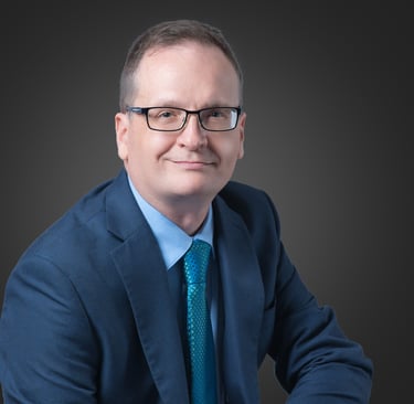 Professional corporate headshot of a smiling businessman wearing glasses, a blue suit, and a teal tie.