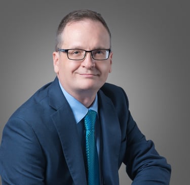 Professional headshot of a businessman in a blue suit and glasses against a grey background.