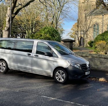 Silver Mercedes-Benz Vito wedding taxi parked outside a traditional stone church with autumn trees.