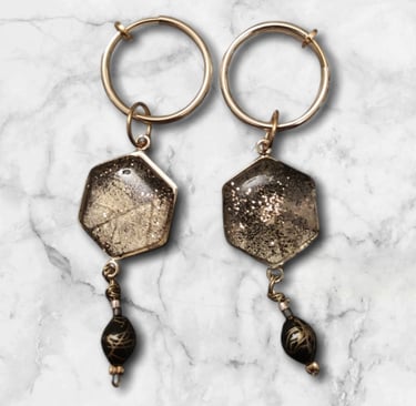 Handmade gold hexagon drop earrings with glitter resin and black beads on a white marble background.