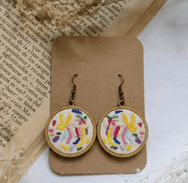 Pendientes de aro con bordado Tenango hecho a mano, con hilos coloridos sobre un fondo vintage.