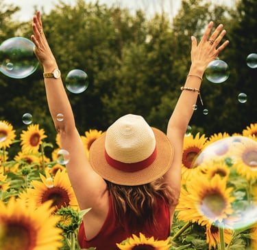 Sommerliche Lebensfreude ab 40: Frau genießt die Sonne im Sonnenblumenfeld mit Seifenblasen.