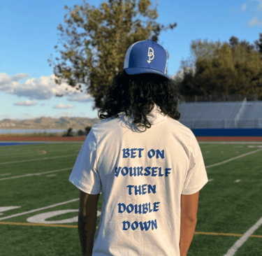 A person wearing a Bet On Yourself graphic t-shirt and blue trucker hat on a football field.