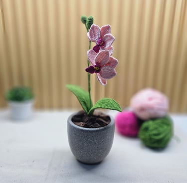 Handmade pink crochet orchid plant in a grey pot with green leaves and yarn balls in the background.