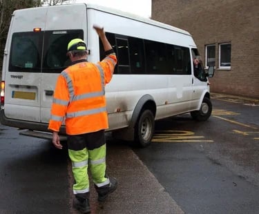A traffic marshal in hi-vis clothing directing a white Ford Transit minibus at a commercial facility.