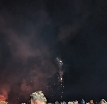 A large crowd of people gathered at night watching a fireworks display and bonfire with smoke in the sky.