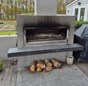 A modern outdoor concrete fireplace on a backyard deck with stacked firewood and a tall chimney.
