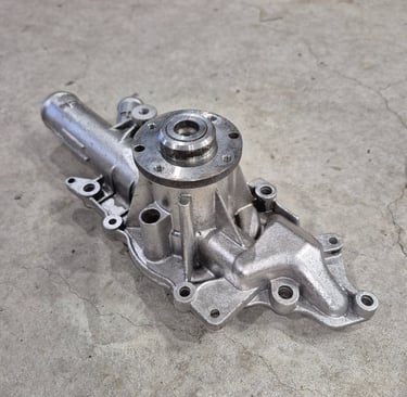 A new metallic automotive engine water pump replacement part sitting on a concrete workshop floor.