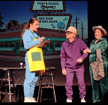 Actors in colorful tracksuits perform a comedic theater scene in front of a retro diner backdrop.