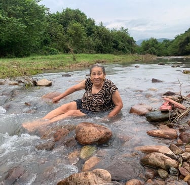 An elderly woman smiling while sitting in a shallow, rocky river surrounded by lush tropical greenery.