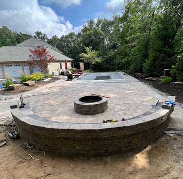 Custom paver patio installation with stone retaining wall, fire pit, and pool deck in a backyard.