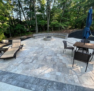 Custom stone paver patio featuring a round fire pit, curved seating wall, lounge chairs, and a dining set.