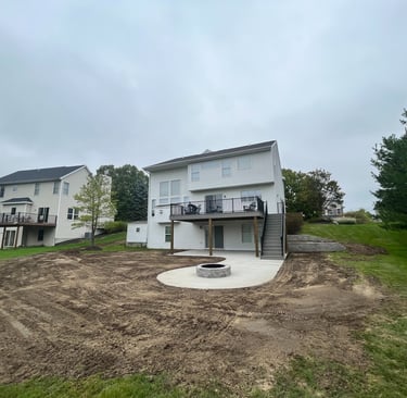 Backyard landscaping project with a new concrete patio, fire pit, and wooden deck under a cloudy sky.
