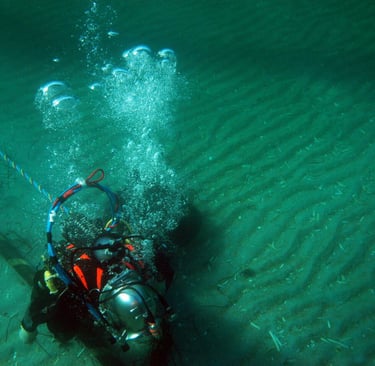 Professional commercial diver in a metal helmet working on an underwater pipeline on the sandy ocean floor.