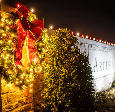A festive Christmas wreath with a red bow and glowing lights on a stone wall entrance to HOA.
