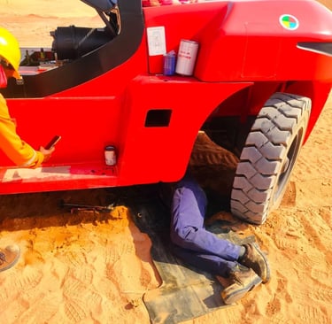 A heavy equipment mechanic performs maintenance under a red industrial forklift in a sandy desert site.