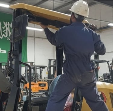 A technician in a hard hat performs forklift maintenance in a Saudi Arabian warehouse facility.