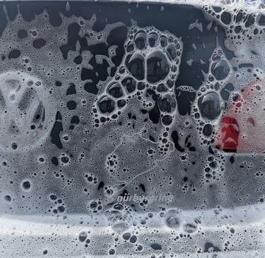 White soapy foam bubbles covering the rear window of a black car during a professional car wash.