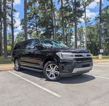 A black Ford Expedition SUV parked in a paved lot surrounded by tall pine trees.