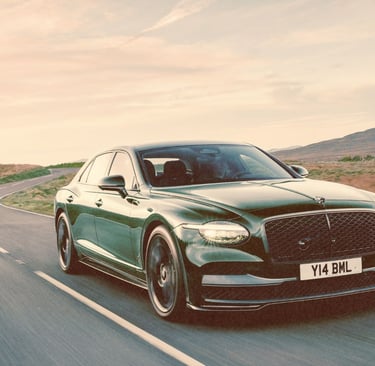 A dark green Bentley Flying Spur luxury sedan driving on a winding countryside road at sunset.