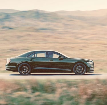 A dark green luxury Bentley Flying Spur sedan driving fast on a scenic mountain road.