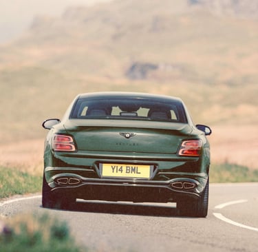 Rear view of a dark green Bentley luxury sedan driving on a winding mountain road.