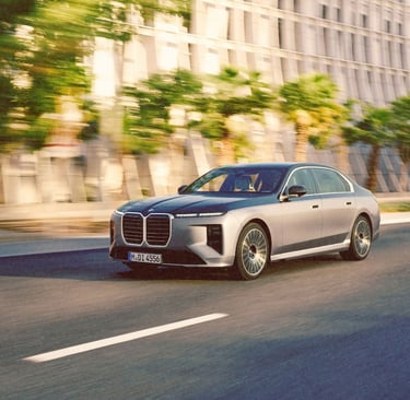 A silver BMW i7 luxury electric sedan driving on a city street past modern architecture.
