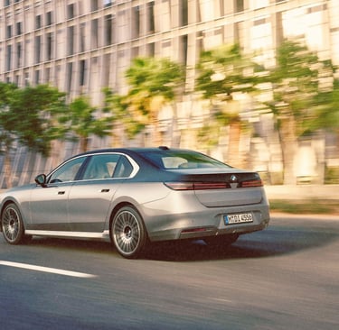 Luxury BMW i7 electric sedan in two-tone grey driving on a modern city street with palm trees.