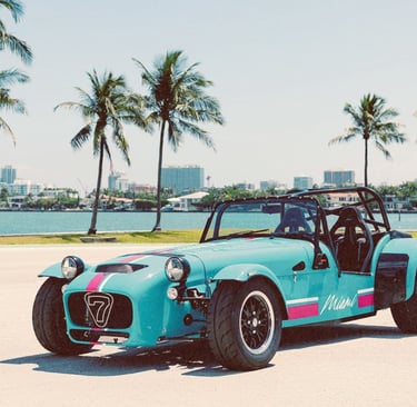 A blue Caterham Seven sportscar parked in Miami with palm trees and a city skyline background.