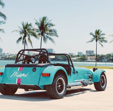 A light blue Caterham sports car parked on a Miami waterfront with tropical palm trees.
