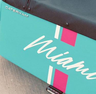 Close-up of a teal Caterham sports car featuring a Miami livery with pink and white stripes.