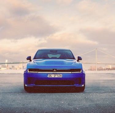 Front view of a blue Dodge Charger electric car parked near a suspension bridge at sunset.