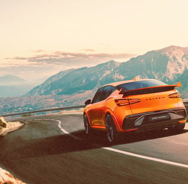 An orange Genesis GV60 Magma performance electric SUV driving on a winding mountain road.