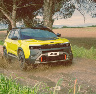 Yellow Jeep Avenger 4xe driving through a mud puddle on a dirt road lined with trees.