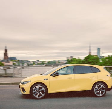 A modern yellow electric hatchback car driving across a city bridge at dusk with a blurred skyline background.