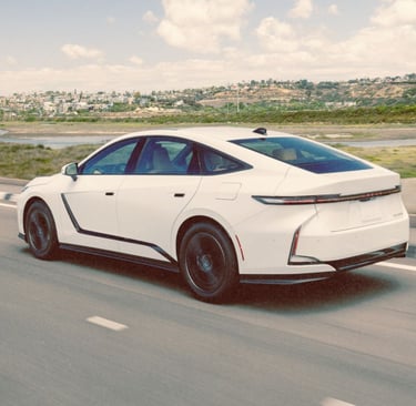 White Lexus ES sedan driving on a coastal road with hills and houses in the background.