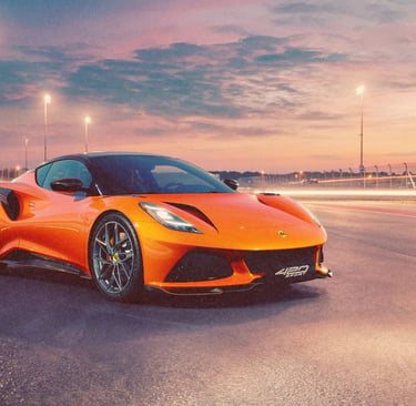 Orange Lotus Emira sports car parked on a race track at sunset with glowing stadium lights.