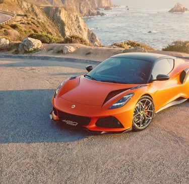 Orange Lotus Emira sports car parked on a scenic coastal cliff road overlooking the Pacific Ocean.