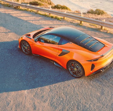 Orange Lotus Emira sports car parked on a scenic coastal road at sunset.