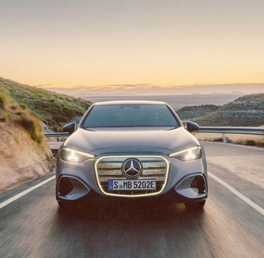A silver Mercedes-Benz electric car driving on a winding coastal road at sunset with glowing LED grille.