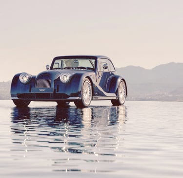 A vintage-style dark blue Morgan sports car parked on a reflective surface with mountain views.