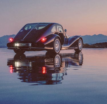 A vintage black luxury coupe reflected in water during a mountain sunset.