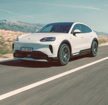 White Porsche Cayenne Electric driving on a coastal road with mountain scenery.