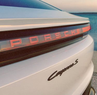 Close-up of the rear taillight and logo of a white Porsche Cayenne S parked by the ocean.