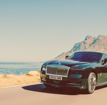 A black Rolls-Royce Spectre luxury electric car driving on a coastal road with mountains.