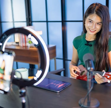 Smiling woman playing ukulele for a live stream with a ring light and smartphone.