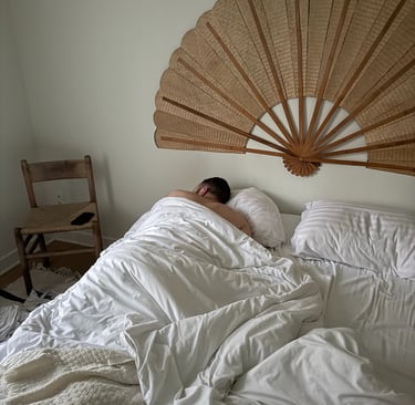 A man sleeping in a bed with white linens below a large decorative rattan fan headboard.