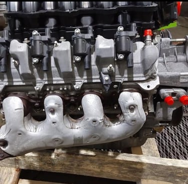A high-performance V8 car engine with silver exhaust manifold and black ignition coils on a wooden pallet.